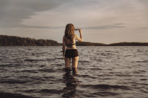 Person står i bikini med vatten upp till knäna och njuter av ett glas rödvin på Friibergh